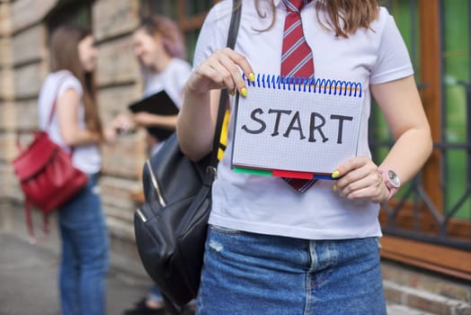 Start. Girl student holds notebook with word start, beginning of classes at school, in college. Brick building and talking students background