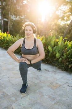 Adorable fat woman in tracksuit is engaged in fitness outdoor side view portrait. Young overweight woman lunges outdoors on warm summer day. Healthy lifestyle and weight loss.