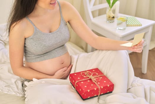 Young beautiful pregnant woman with gift box, female sitting at home in bed reading a greeting card