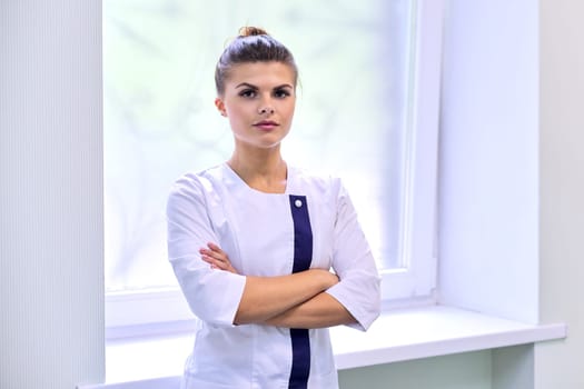 Portrait of young attractive confident woman in medical uniform with arms crossed, light wall clinic window background. Female pharmacist, nurse, cosmetologist, specialist, scientist