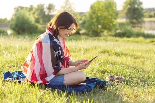 Adult woman sitting on July evening in nature with USA flag scarf, female in headphones with smartphone looking Independence Day celebration