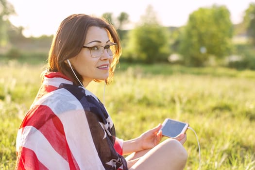 Adult woman sitting on July evening in nature with USA flag scarf, female in headphones with smartphone looking Independence Day celebration