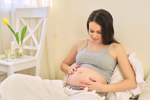 Young beautiful pregnant woman sitting at home in bed, female touching her tummy, waiting for the birth of baby