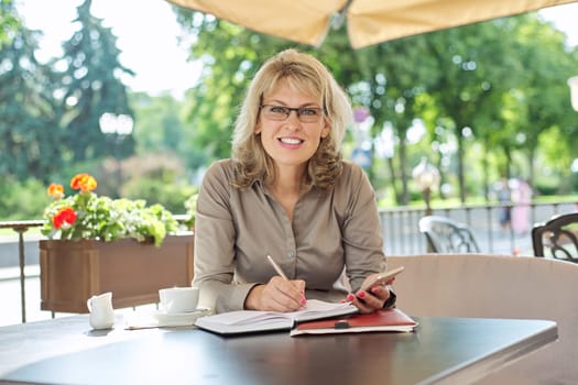 Business beautiful woman writing in business notebook using smartphone, smiling female sitting in summer outdoor cafe, coffee break