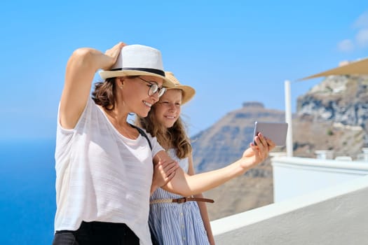 Happy tourists mother and daughter child taking selfie photo on smartphone while traveling around Santorini island. Family trip, famous travel destinations
