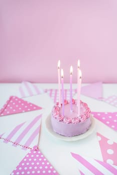 Delicate lavender pink bento cake on a plate on a pink background of party flags, candles burning on the cake. Celebration, happy day