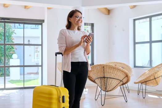 Woman tourist hotel guest with suitcase in the lobby of hotel using smartphone. Travelling, vacation, leisure, business trip, people