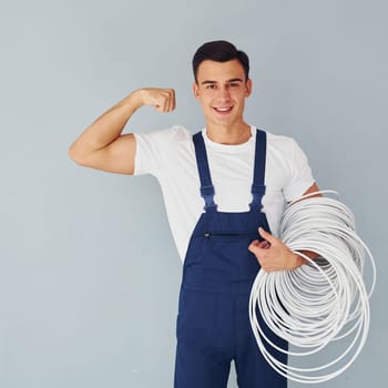 Holds cable. Male worker in blue uniform standing inside of studio against white background.