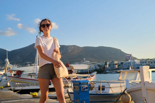 Beautiful fashionable girl teenager walking along sea promenade, pier with yachts. Mountains, sunset on sea background, copy space. Leisure, resort, beauty, youth, fashion, young people concept