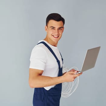 Uses laptop. Male worker in blue uniform standing inside of studio against white background.