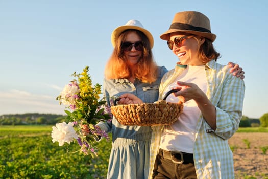 Portrait of two happy smiling embraced women mother and teenage daughter with basket of farm eggs in hats, with bouquet of flowers in garden, summer nature, sunset background