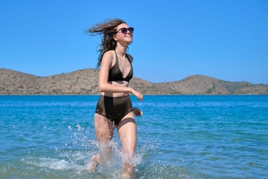 Beautiful laughing teenager girl in sunglasses swimsuit bikini running on sea water, sunny summer day. Relax, fun, vacation on the beach