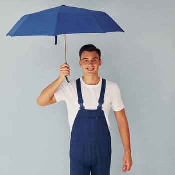 Holds umbrella by hand. Male worker in blue uniform standing inside of studio against white background.