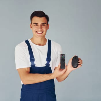 TV remote controller and computer mouse. Male worker in blue uniform standing inside of studio against white background.