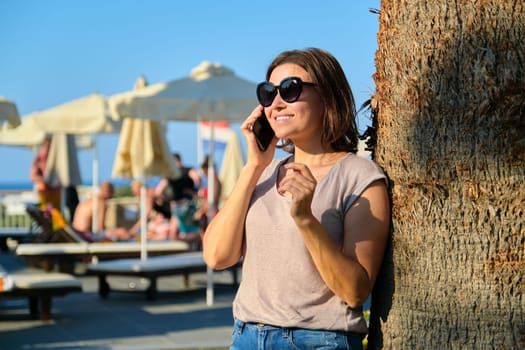 Portrait of beautiful smiling mature woman relaxing in resort hotel, female in sunglasses standing near palm tree using smartphone, copy space. Vacation, summer, tourism, resort, middle-aged people