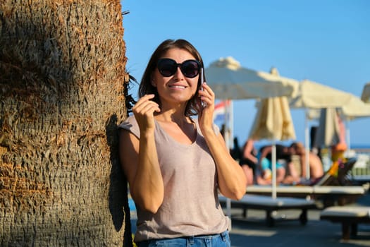 Portrait of beautiful smiling mature woman relaxing in resort hotel, female in sunglasses standing near palm tree using smartphone, copy space. Vacation, summer, tourism, resort, middle-aged people