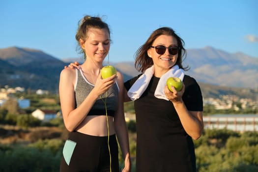 Healthy lifestyle and healthy food concept. Smiling fitness mother and teen daughter together eating apples, talking, resting after athletic running and outdoor exercise