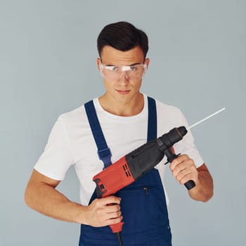 In protective eyewear and with drill in hands. Male worker in blue uniform standing inside of studio against white background.