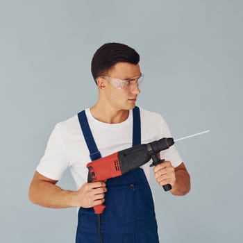 In protective eyewear and with drill in hands. Male worker in blue uniform standing inside of studio against white background.