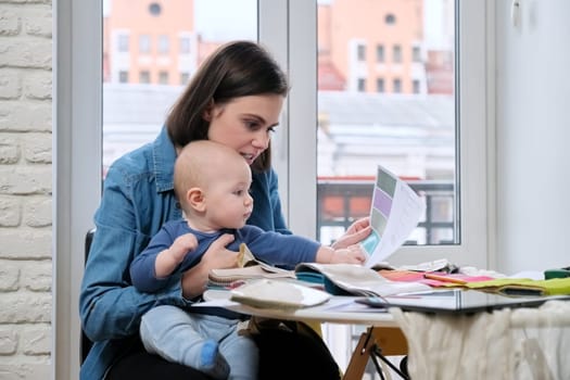 Young mother work with baby in her arms at home office. Woman interior designer working with fabric samples, on the table tablet computer, sketches and textile pallets