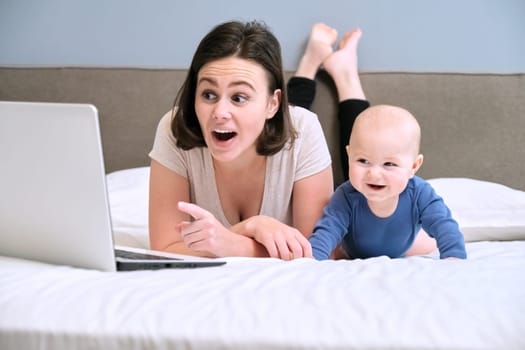 Happy smiling mother and baby son lying together at home on bed and look at laptop computer, work and leisure of young mother