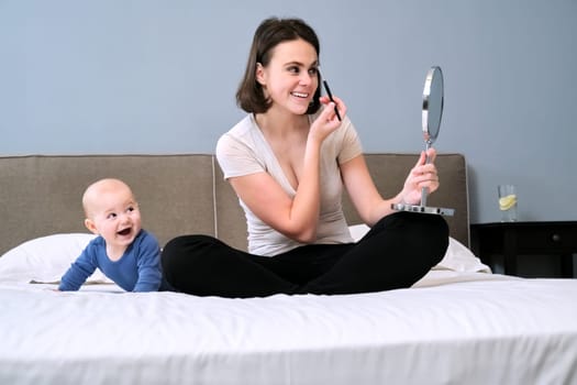 Young beautiful happy mother with toddler son at home in bed, woman with cosmetic mirror does makeup