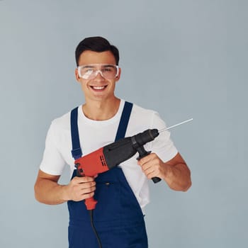 Holds drill in hands. Male worker in blue uniform standing inside of studio against white background.