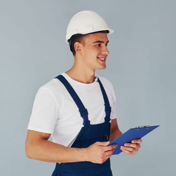 With notepad and hard hat. Male worker in blue uniform standing inside of studio against white background.