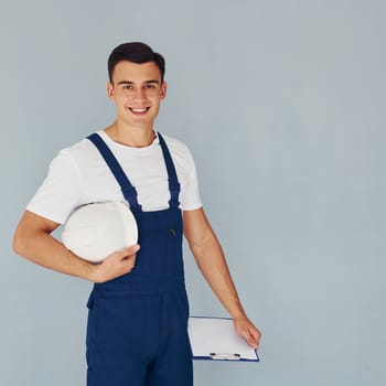 With notepad and hard hat. Male worker in blue uniform standing inside of studio against white background.