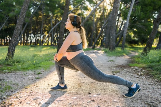 Adorable fat woman in tracksuit is engaged in fitness outdoor side view portrait. Young overweight woman lunges outdoors on warm summer day. Healthy lifestyle and weight loss.