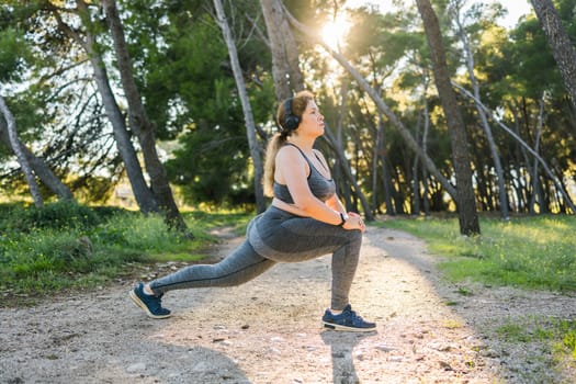 Adorable fat woman in tracksuit is engaged in fitness outdoor side view portrait. Young overweight woman lunges outdoors on warm summer day. Healthy lifestyle and weight loss.