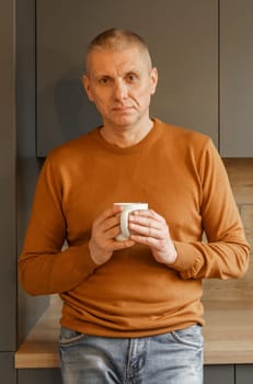 Portrait of a mature man in an orange jumper in the kitchen with a mug of warm tea. Vertical frame.