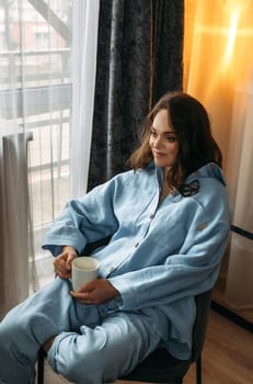 Portrait of a young woman in a light blue suit, who is sitting on a chair with a white cup in her hands. Vertical frame.