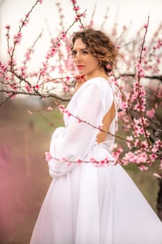 Woman peach blossom. Happy curly woman in white dress walking in the garden of blossoming peach trees in spring.