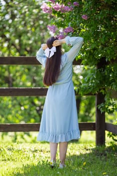 brunette girl with long hair with white bow, in light blue dress, stands in the garden, nearby blooming lilac bush , in sunny day. Back view. Close up. Vertical. copy space