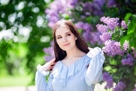 Pretty brunette girl with long hair stands with lilac flowers, in the garden, in sunny day. Close up. copy space