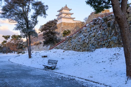 Historic Japanese castle towers over snow covered bench along path in park. High quality photo