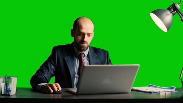 Businessman working on laptop over greenscreen backdrop, using documents at desk. Successful team leader in suit posing over isolated chroma key template, blank copyspace background.