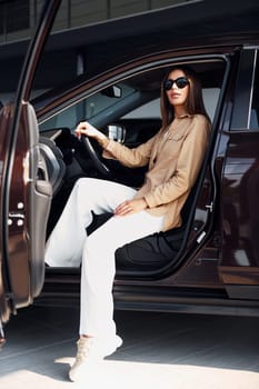 Sits with car's window opened. Fashionable beautiful young woman and her modern automobile.