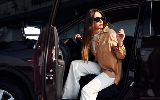 Sits with car's window opened. Fashionable beautiful young woman and her modern automobile.