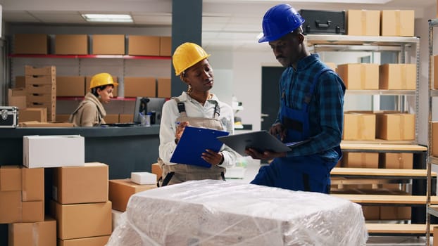 African american people in overalls analyzing products placed on storage room shelves, cargo shipping in cardboard boxes. Man and woman working with industrial goods, depot space.