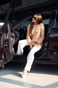 Sits with car's window opened. Fashionable beautiful young woman and her modern automobile.