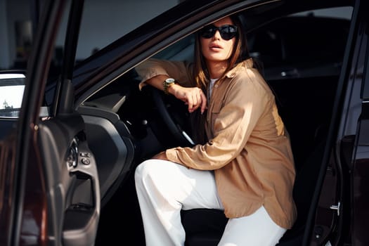 Sits with car's window opened. Fashionable beautiful young woman and her modern automobile.