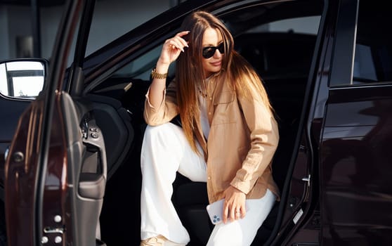 Sits with car's window opened. Fashionable beautiful young woman and her modern automobile.