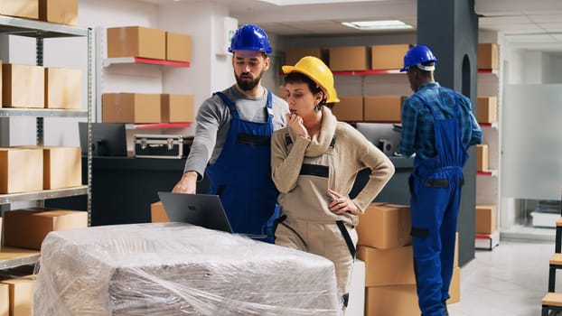 Man and woman thinking about packing goods in boxes, looking at stock logistics on laptop and counting products. Warehouse employee and supervisor with overalls and hardhat, delivery service.