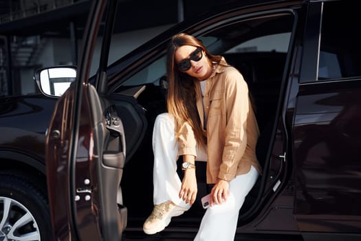 Sits with car's window opened. Fashionable beautiful young woman and her modern automobile.