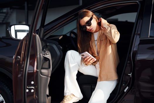 Sits with car's window opened. Fashionable beautiful young woman and her modern automobile.