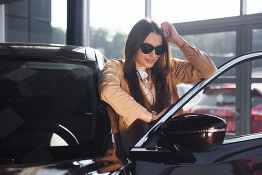 Standing near opened window. Fashionable beautiful young woman and her modern automobile.