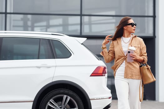 Outdoors against modern building. Fashionable beautiful young woman and her modern automobile.