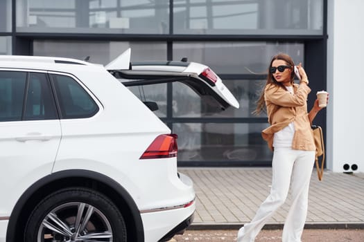 Outdoors against modern building. Fashionable beautiful young woman and her modern automobile.